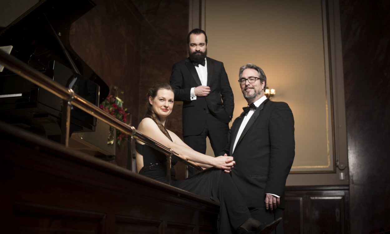 Drei elegant gekleidete Musikerinnen und Musiker in Abendgarderobe posieren neben einem Flügel in einem Konzertsaal mit warmem Licht.