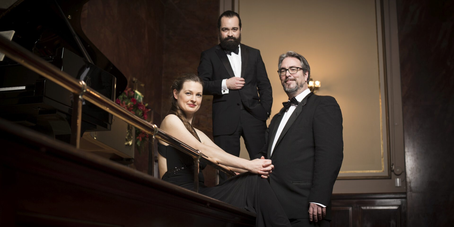 Drei elegant gekleidete Musikerinnen und Musiker in Abendgarderobe posieren neben einem Flügel in einem Konzertsaal mit warmem Licht.