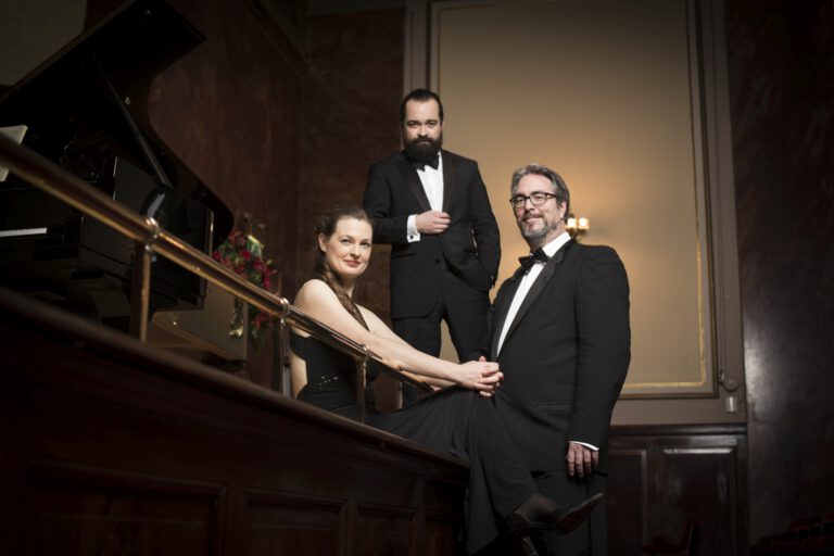 Drei elegant gekleidete Musikerinnen und Musiker in Abendgarderobe posieren neben einem Flügel in einem Konzertsaal mit warmem Licht.