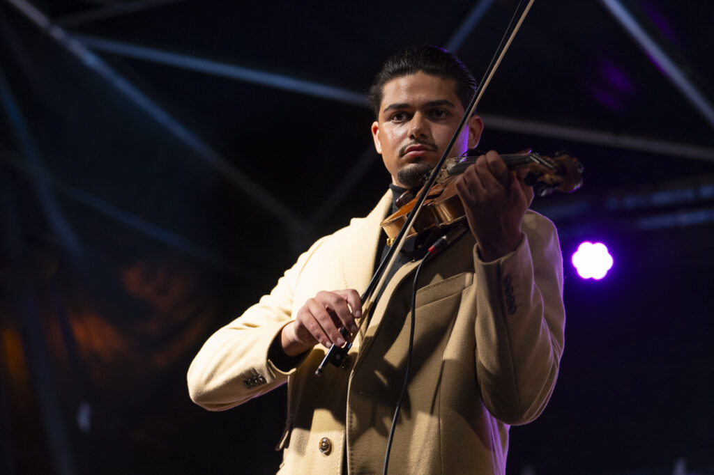 Geiger in hellem Mantel spielt auf einer Bühne Violine, im Hintergrund farbige Scheinwerfer und dunkle Konzertkulisse.