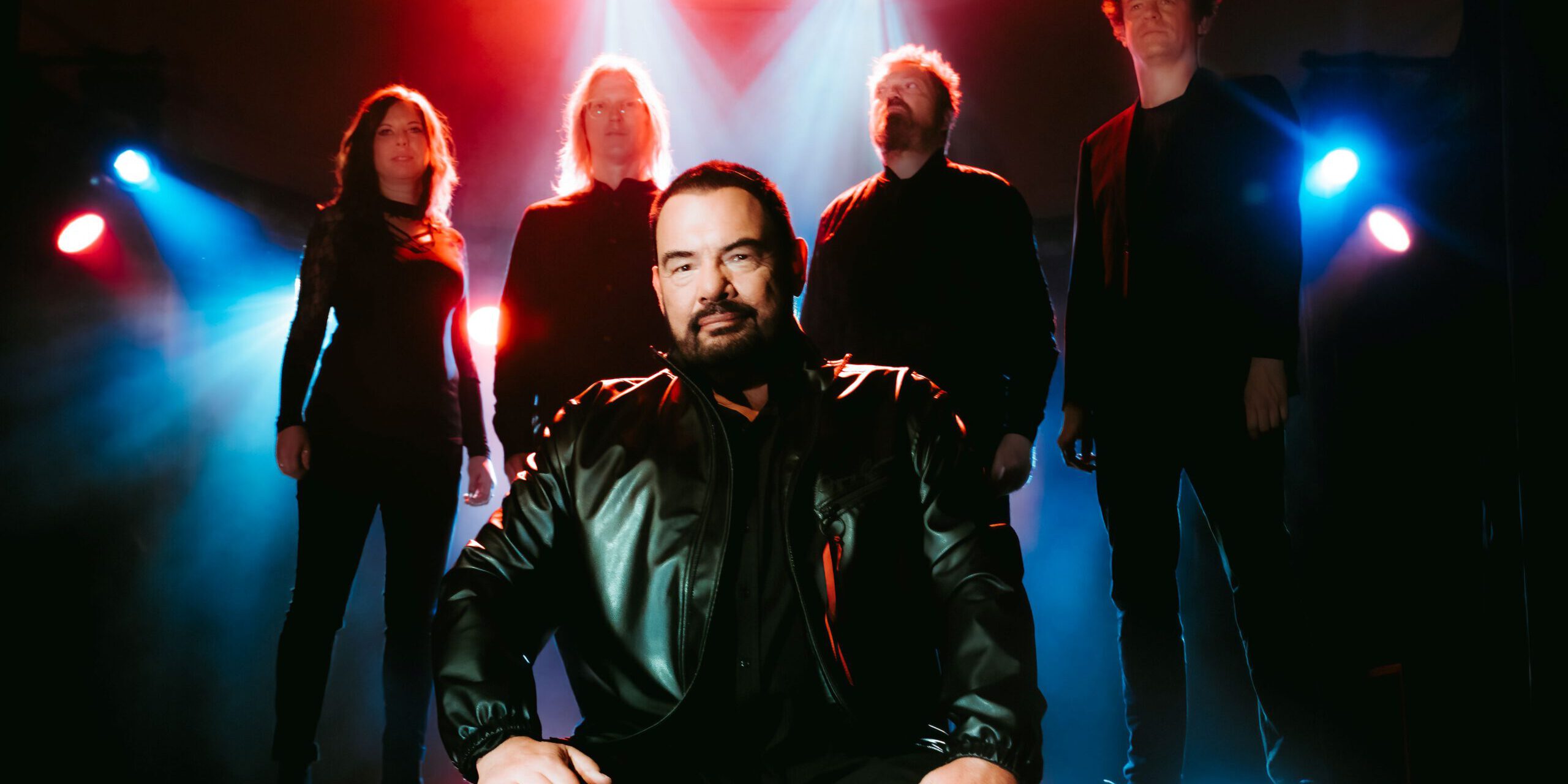 Eine Musikband steht auf einer Bühne im farbigen Bühnenlicht. Im Vordergrund sitzt ein Mann in schwarzer Lederjacke, hinter ihm stehen vier weitere Bandmitglieder in dunkler Kleidung, dramatisch von roten und blauen Scheinwerfern beleuchtet.