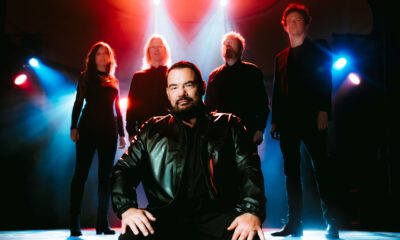 Eine Musikband steht auf einer Bühne im farbigen Bühnenlicht. Im Vordergrund sitzt ein Mann in schwarzer Lederjacke, hinter ihm stehen vier weitere Bandmitglieder in dunkler Kleidung, dramatisch von roten und blauen Scheinwerfern beleuchtet.