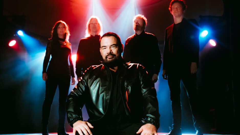 Eine Musikband steht auf einer Bühne im farbigen Bühnenlicht. Im Vordergrund sitzt ein Mann in schwarzer Lederjacke, hinter ihm stehen vier weitere Bandmitglieder in dunkler Kleidung, dramatisch von roten und blauen Scheinwerfern beleuchtet.