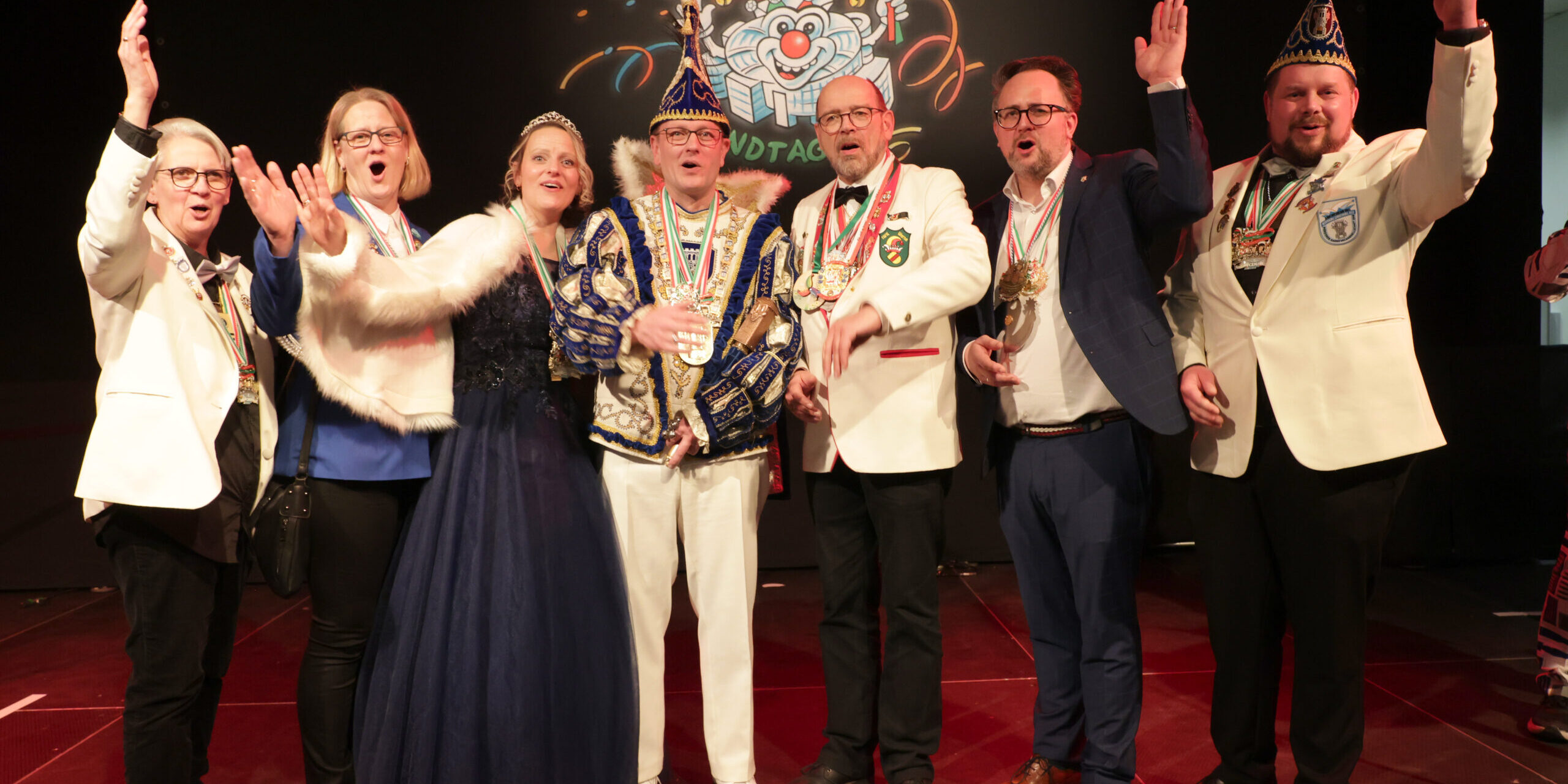 Acht festlich gekleidete Erwachsene stehen auf einer Bühne und jubeln gemeinsam. Einige tragen Karnevalsorden und Narrenkappen, im Hintergrund ist ein bunt beleuchtetes Karnevalsmotiv mit der Aufschrift „Närrischer Landtag“ zu sehen.