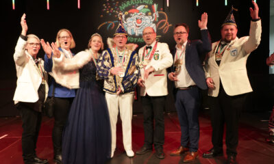 Acht festlich gekleidete Erwachsene stehen auf einer Bühne und jubeln gemeinsam. Einige tragen Karnevalsorden und Narrenkappen, im Hintergrund ist ein bunt beleuchtetes Karnevalsmotiv mit der Aufschrift „Närrischer Landtag“ zu sehen.