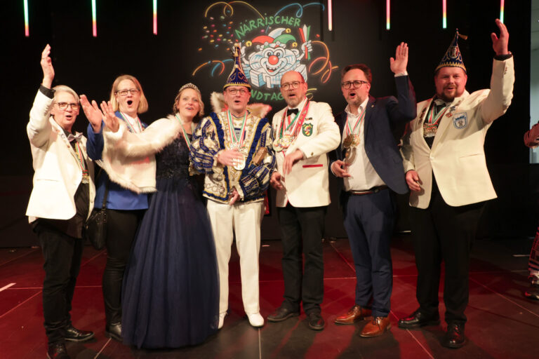 Foto: Bernd Schälte/Landtag NRW Acht festlich gekleidete Erwachsene stehen auf einer Bühne und jubeln gemeinsam. Einige tragen Karnevalsorden und Narrenkappen, im Hintergrund ist ein bunt beleuchtetes Karnevalsmotiv mit der Aufschrift „Närrischer Landtag“ zu sehen.