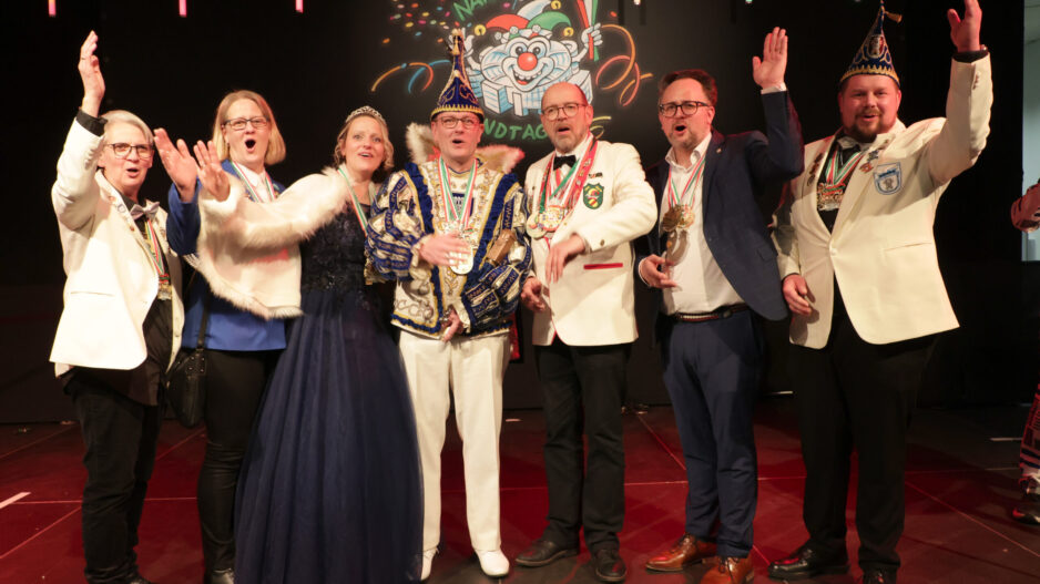 Acht festlich gekleidete Erwachsene stehen auf einer Bühne und jubeln gemeinsam. Einige tragen Karnevalsorden und Narrenkappen, im Hintergrund ist ein bunt beleuchtetes Karnevalsmotiv mit der Aufschrift „Närrischer Landtag“ zu sehen.