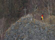 Auf einem felsigen Hügel steht ein schlichtes Kreuz, daneben weht eine deutsche Flagge. Im Hintergrund sind kahle Bäume eines Waldes zu sehen.