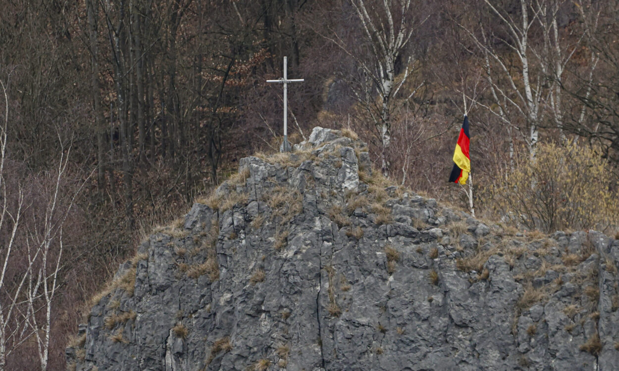 Auf einem felsigen Hügel steht ein schlichtes Kreuz, daneben weht eine deutsche Flagge. Im Hintergrund sind kahle Bäume eines Waldes zu sehen.