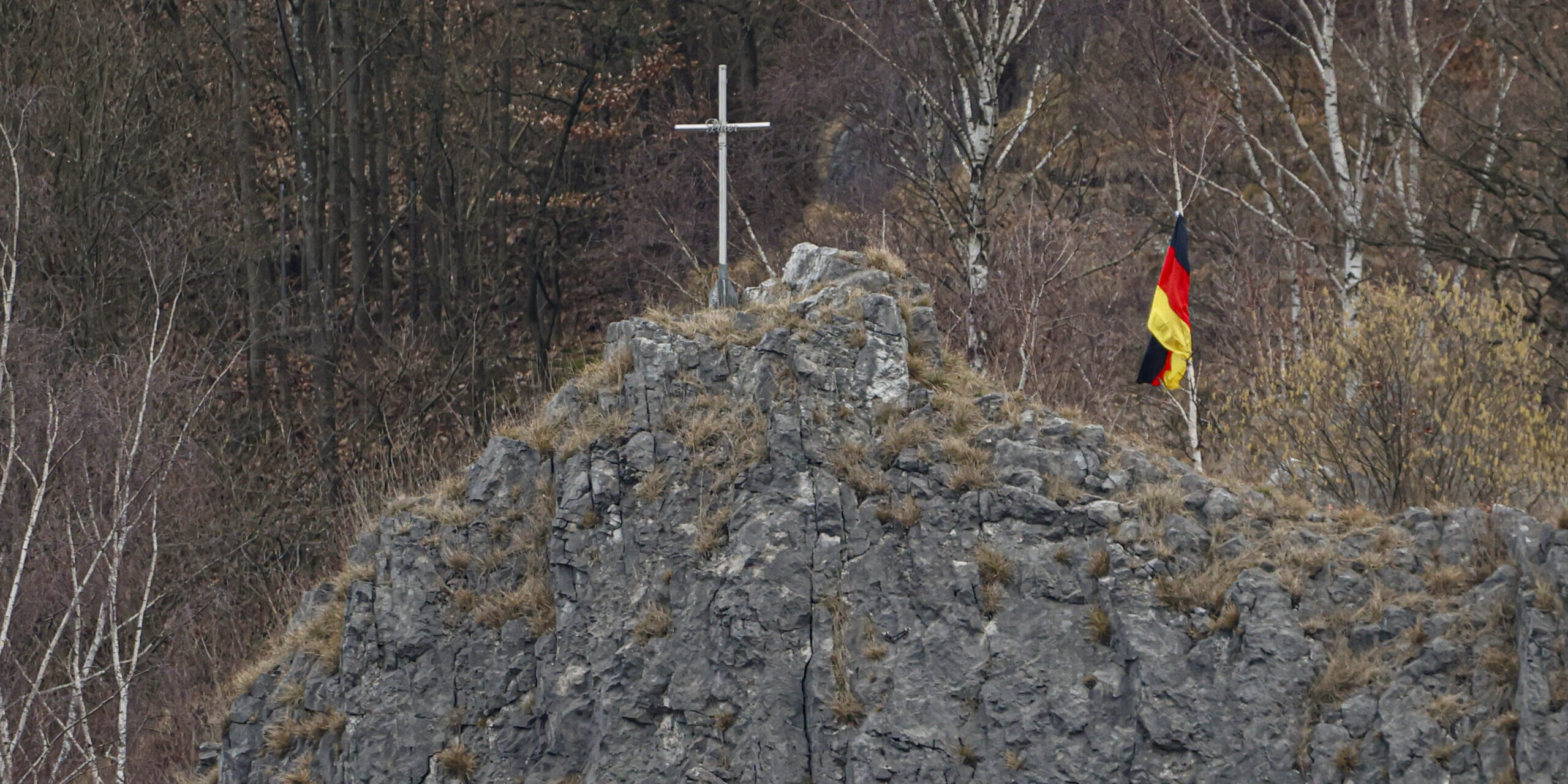 Auf einem felsigen Hügel steht ein schlichtes Kreuz, daneben weht eine deutsche Flagge. Im Hintergrund sind kahle Bäume eines Waldes zu sehen.