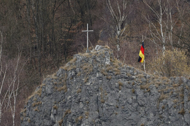 Auf einem felsigen Hügel steht ein schlichtes Kreuz, daneben weht eine deutsche Flagge. Im Hintergrund sind kahle Bäume eines Waldes zu sehen.