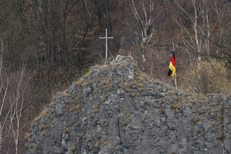 Auf einem felsigen Hügel steht ein schlichtes Kreuz, daneben weht eine deutsche Flagge. Im Hintergrund sind kahle Bäume eines Waldes zu sehen.