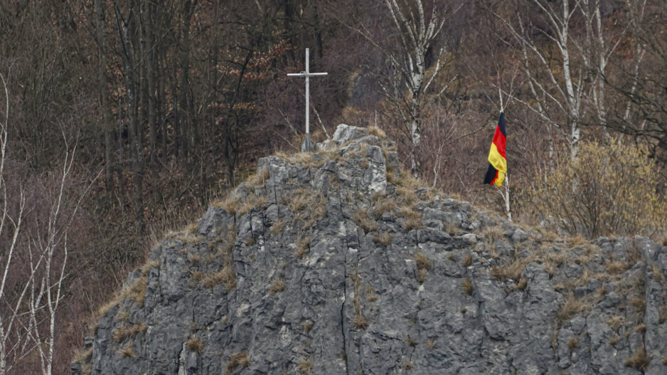 Auf einem felsigen Hügel steht ein schlichtes Kreuz, daneben weht eine deutsche Flagge. Im Hintergrund sind kahle Bäume eines Waldes zu sehen.
