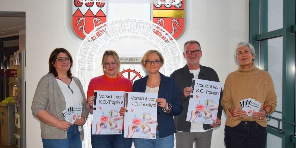Fünf Erwachsene stehen nebeneinander in einem Innenraum und halten Plakate mit der Aufschrift „Vorsicht vor K.O.-Tropfen!“. Hinter ihnen ist eine leuchtende Installation sowie ein Stadtwappen an der Wand zu sehen.