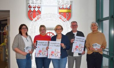 Fünf Erwachsene stehen nebeneinander in einem Innenraum und halten Plakate mit der Aufschrift „Vorsicht vor K.O.-Tropfen!“. Hinter ihnen ist eine leuchtende Installation sowie ein Stadtwappen an der Wand zu sehen.