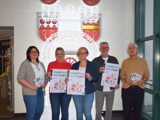 Fünf Erwachsene stehen nebeneinander in einem Innenraum und halten Plakate mit der Aufschrift „Vorsicht vor K.O.-Tropfen!“. Hinter ihnen ist eine leuchtende Installation sowie ein Stadtwappen an der Wand zu sehen.
