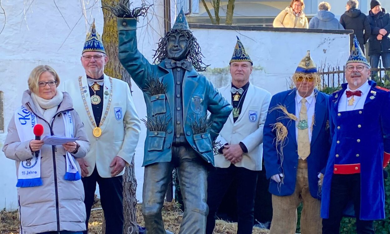 Mehrere festlich gekleidete Erwachsene stehen im Freien neben einer bronzenen Statue. Einige tragen Karnevalsmützen und Orden, eine Frau hält ein Mikrofon und liest von einem Blatt. Im Hintergrund sind eine Mauer, Bäume und Zuschauer zu sehen.