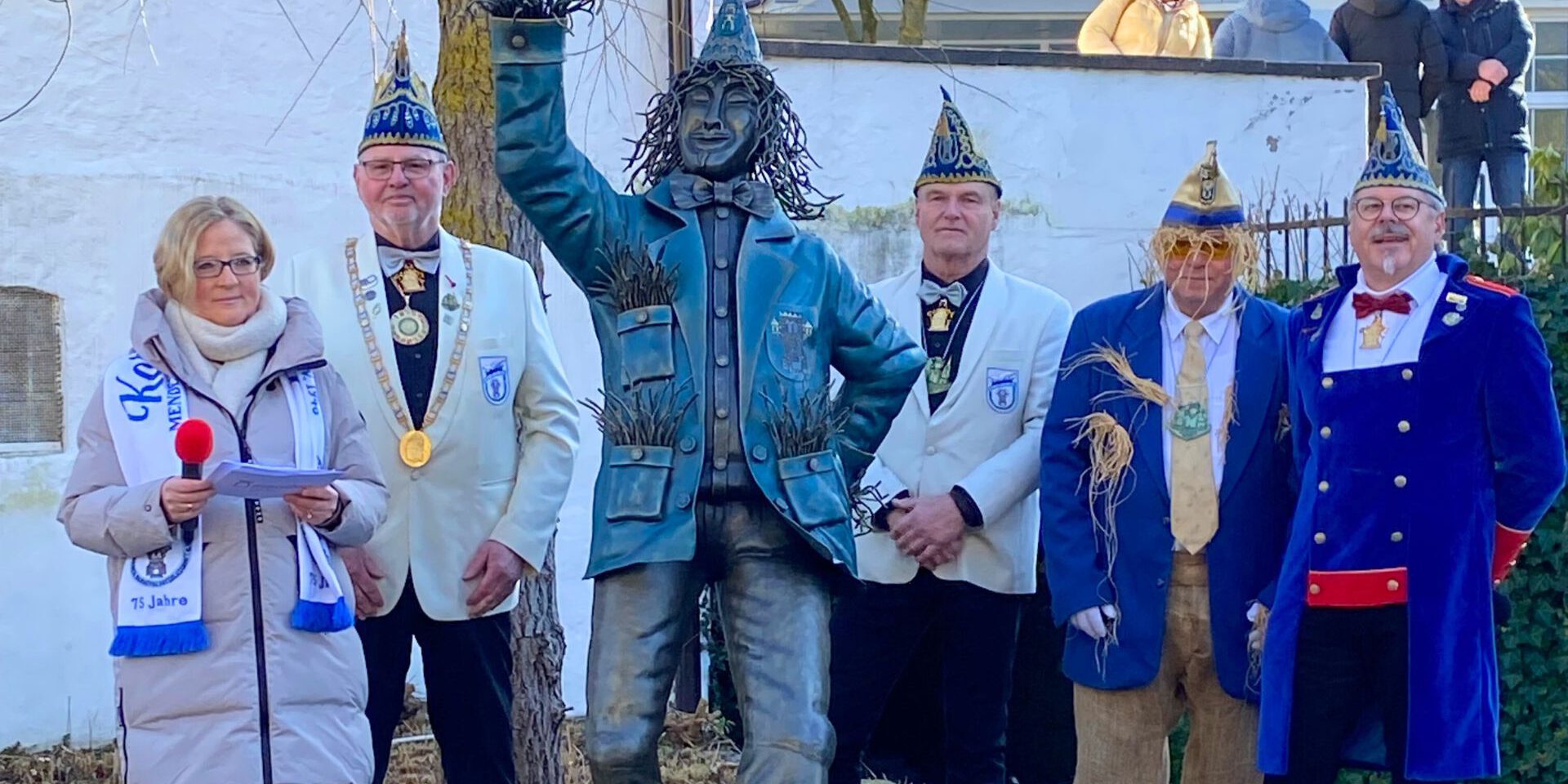 Mehrere festlich gekleidete Erwachsene stehen im Freien neben einer bronzenen Statue. Einige tragen Karnevalsmützen und Orden, eine Frau hält ein Mikrofon und liest von einem Blatt. Im Hintergrund sind eine Mauer, Bäume und Zuschauer zu sehen.