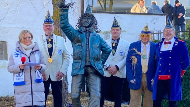Mehrere festlich gekleidete Erwachsene stehen im Freien neben einer bronzenen Statue. Einige tragen Karnevalsmützen und Orden, eine Frau hält ein Mikrofon und liest von einem Blatt. Im Hintergrund sind eine Mauer, Bäume und Zuschauer zu sehen.