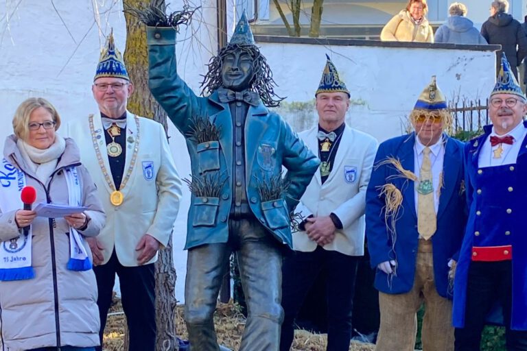 Mehrere festlich gekleidete Erwachsene stehen im Freien neben einer bronzenen Statue. Einige tragen Karnevalsmützen und Orden, eine Frau hält ein Mikrofon und liest von einem Blatt. Im Hintergrund sind eine Mauer, Bäume und Zuschauer zu sehen.