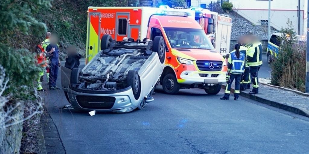 Ein umgestürzter Pkw liegt auf dem Dach auf einer Straße. Mehrere Rettungskräfte stehen um das Fahrzeug, dahinter sind ein Rettungswagen und weitere Einsatzfahrzeuge mit Blaulicht zu sehen.