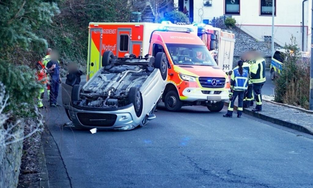 Ein umgestürzter Pkw liegt auf dem Dach auf einer Straße. Mehrere Rettungskräfte stehen um das Fahrzeug, dahinter sind ein Rettungswagen und weitere Einsatzfahrzeuge mit Blaulicht zu sehen.