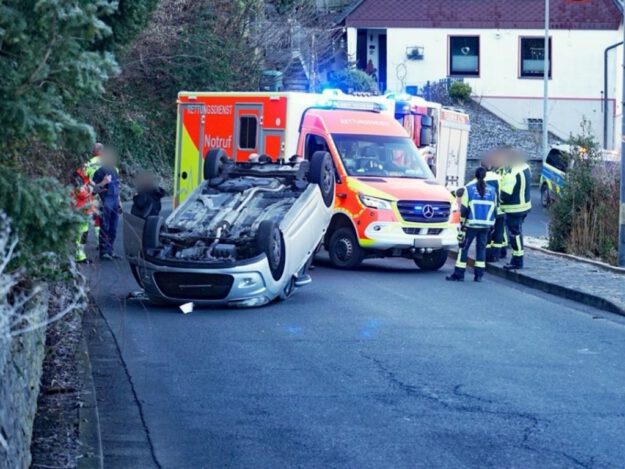 Ein umgestürzter Pkw liegt auf dem Dach auf einer Straße. Mehrere Rettungskräfte stehen um das Fahrzeug, dahinter sind ein Rettungswagen und weitere Einsatzfahrzeuge mit Blaulicht zu sehen.