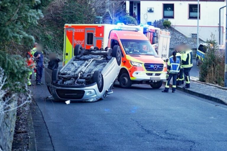 Ein umgestürzter Pkw liegt auf dem Dach auf einer Straße. Mehrere Rettungskräfte stehen um das Fahrzeug, dahinter sind ein Rettungswagen und weitere Einsatzfahrzeuge mit Blaulicht zu sehen.
