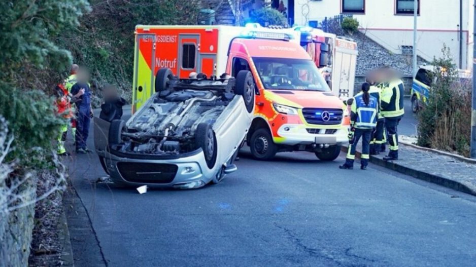 Ein umgestürzter Pkw liegt auf dem Dach auf einer Straße. Mehrere Rettungskräfte stehen um das Fahrzeug, dahinter sind ein Rettungswagen und weitere Einsatzfahrzeuge mit Blaulicht zu sehen.
