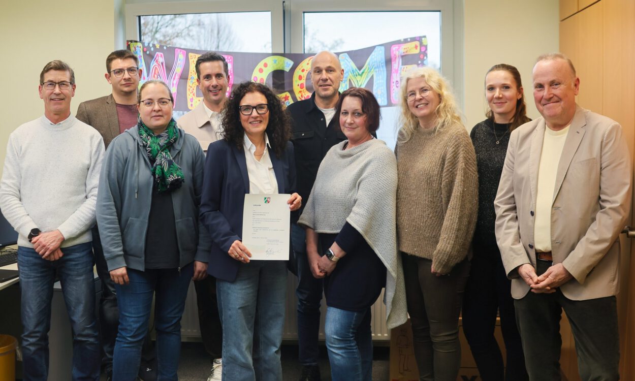 Gruppe von zehn Personen steht in einem Büro vor einem „Welcome“-Schild; eine Frau in der Mitte hält ein offizielles Dokument in die Kamera.