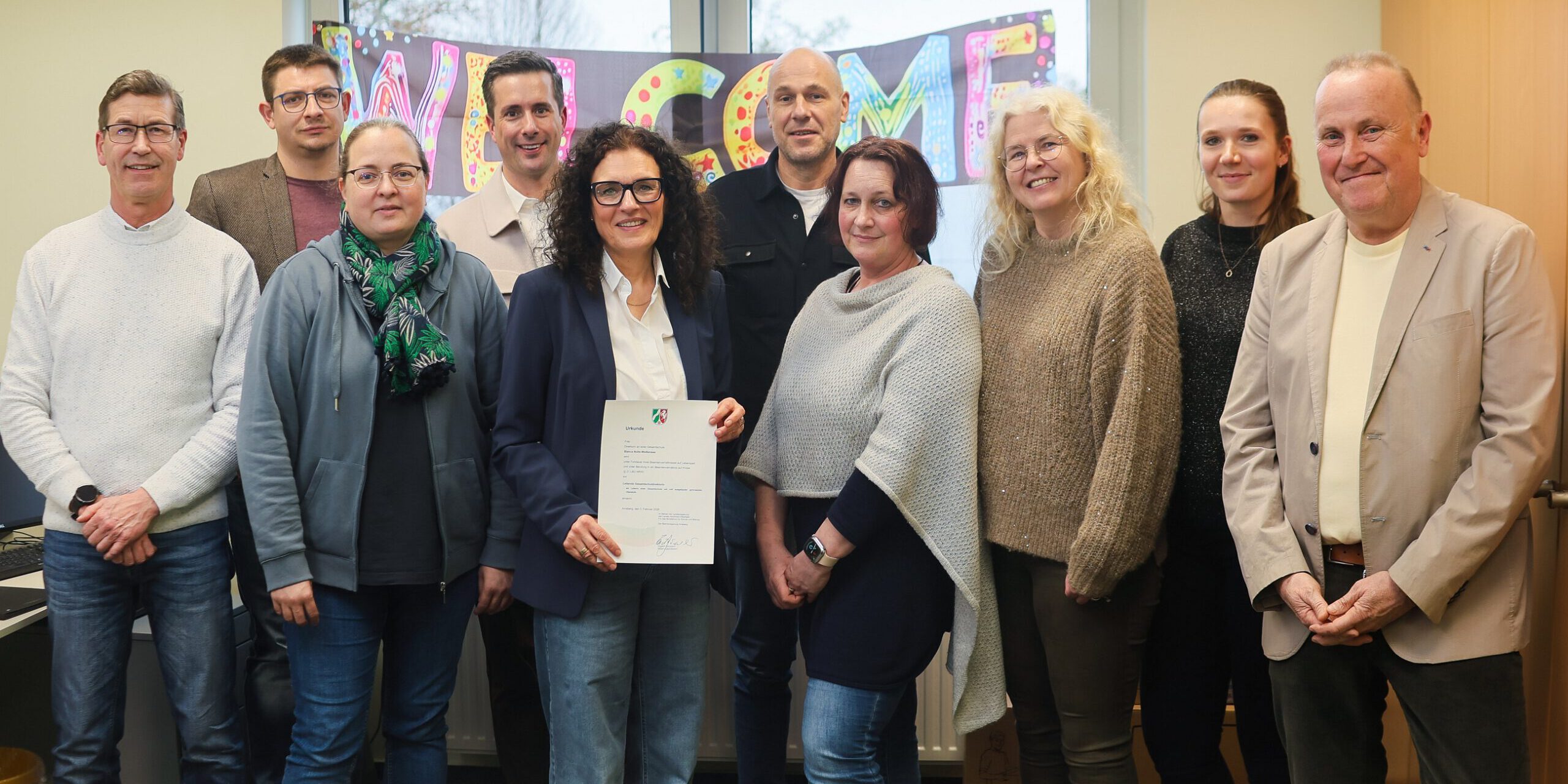 Gruppe von zehn Personen steht in einem Büro vor einem „Welcome“-Schild; eine Frau in der Mitte hält ein offizielles Dokument in die Kamera.
