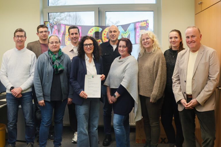 Gruppe von zehn Personen steht in einem Büro vor einem „Welcome“-Schild; eine Frau in der Mitte hält ein offizielles Dokument in die Kamera.