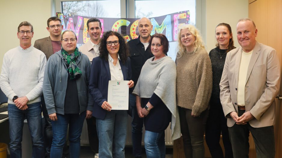 Gruppe von zehn Personen steht in einem Büro vor einem „Welcome“-Schild; eine Frau in der Mitte hält ein offizielles Dokument in die Kamera.