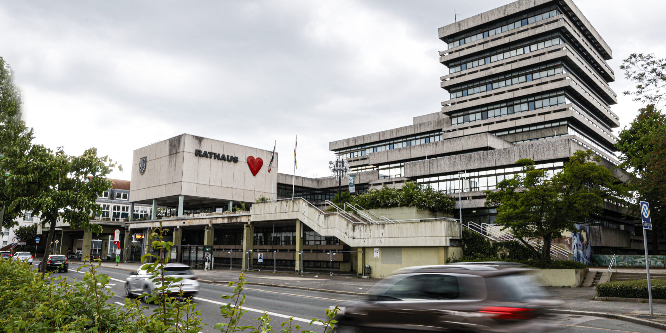 Modernes Rathausgebäude mit gestaffeltem Hochhaus und „Rathaus“-Schriftzug samt rotem Herz an der Fassade; davor eine Straße mit vorbeifahrenden Autos unter bewölktem Himmel.