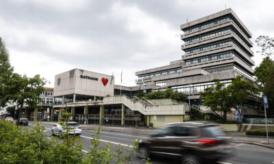 Modernes Rathausgebäude mit gestaffeltem Hochhaus und „Rathaus“-Schriftzug samt rotem Herz an der Fassade; davor eine Straße mit vorbeifahrenden Autos unter bewölktem Himmel.