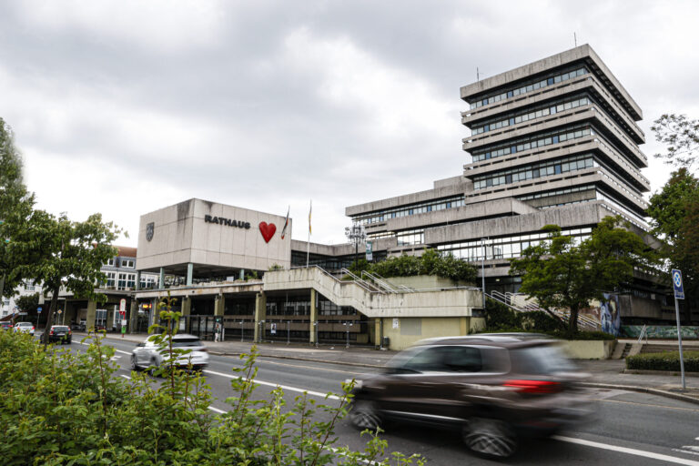 Modernes Rathausgebäude mit gestaffeltem Hochhaus und „Rathaus“-Schriftzug samt rotem Herz an der Fassade; davor eine Straße mit vorbeifahrenden Autos unter bewölktem Himmel.
