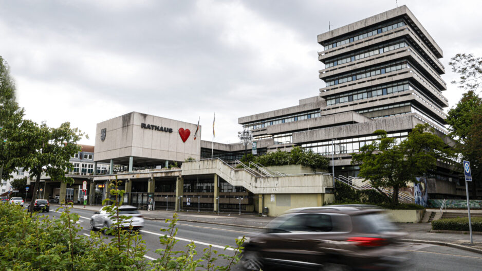 Modernes Rathausgebäude mit gestaffeltem Hochhaus und „Rathaus“-Schriftzug samt rotem Herz an der Fassade; davor eine Straße mit vorbeifahrenden Autos unter bewölktem Himmel.
