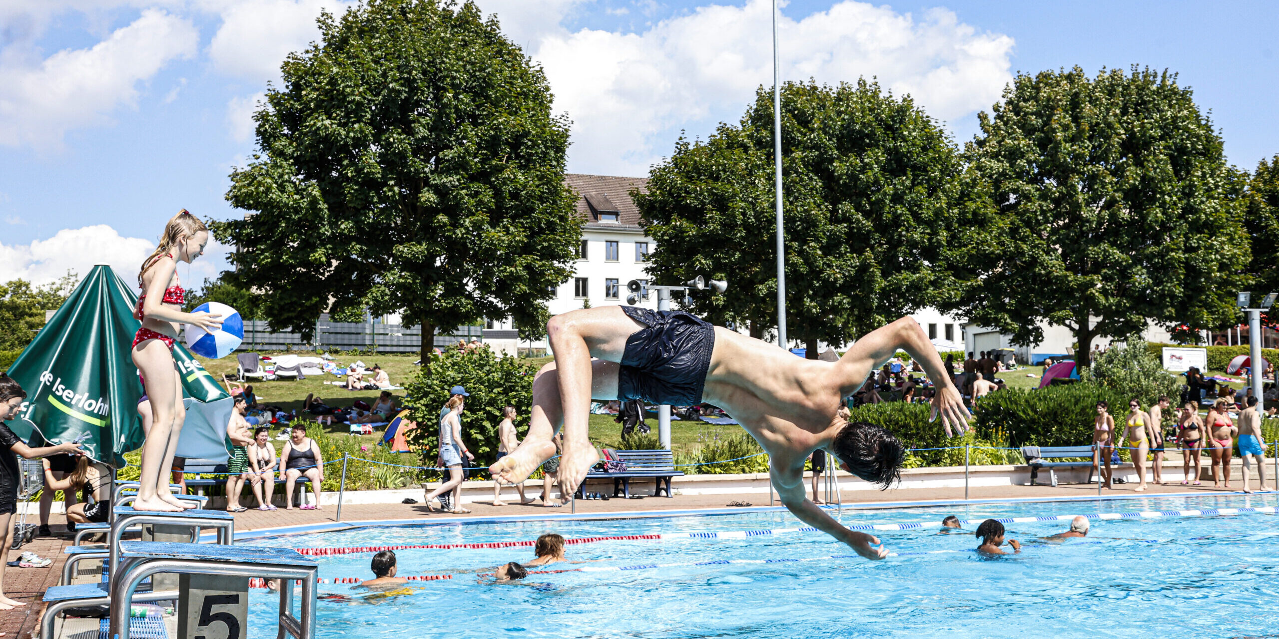 Jugendlicher springt kopfüber von einem Startblock ins Freibadbecken, während andere Badegäste im Wasser schwimmen und am Beckenrand stehen.
