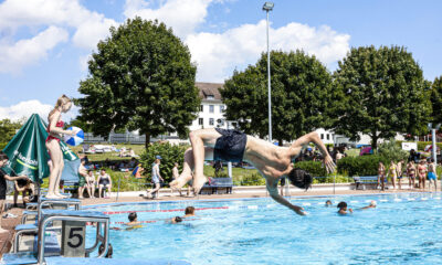 Jugendlicher springt kopfüber von einem Startblock ins Freibadbecken, während andere Badegäste im Wasser schwimmen und am Beckenrand stehen.