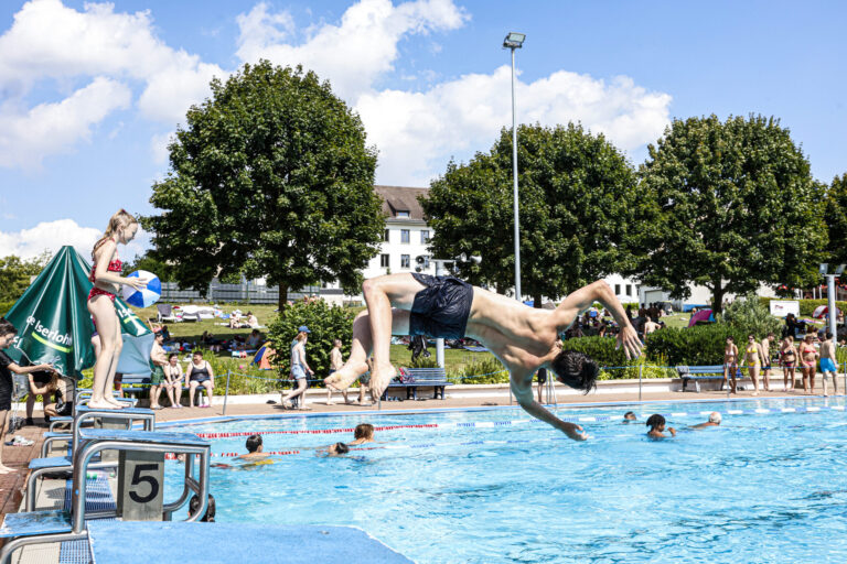 Jugendlicher springt kopfüber von einem Startblock ins Freibadbecken, während andere Badegäste im Wasser schwimmen und am Beckenrand stehen.