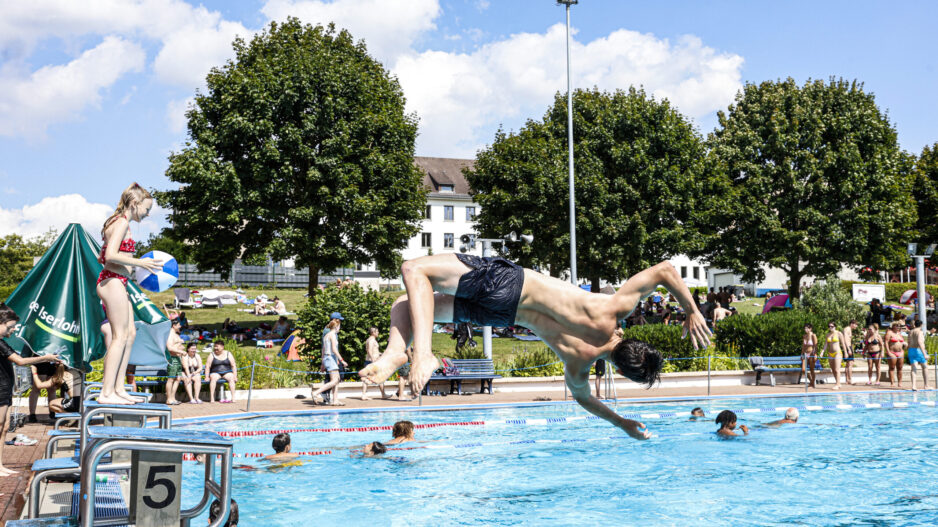 Jugendlicher springt kopfüber von einem Startblock ins Freibadbecken, während andere Badegäste im Wasser schwimmen und am Beckenrand stehen.