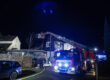 Ein Feuerwehreinsatz im Dunkeln in einem Wohngebiet: Ein Feuerwehrfahrzeug mit ausgefahrener Drehleiter steht vor einem Haus, mehrere Einsatzkräfte arbeiten unter blauem Licht. Schläuche liegen auf der Straße, umliegende Gebäude und parkende Autos sind zu sehen.