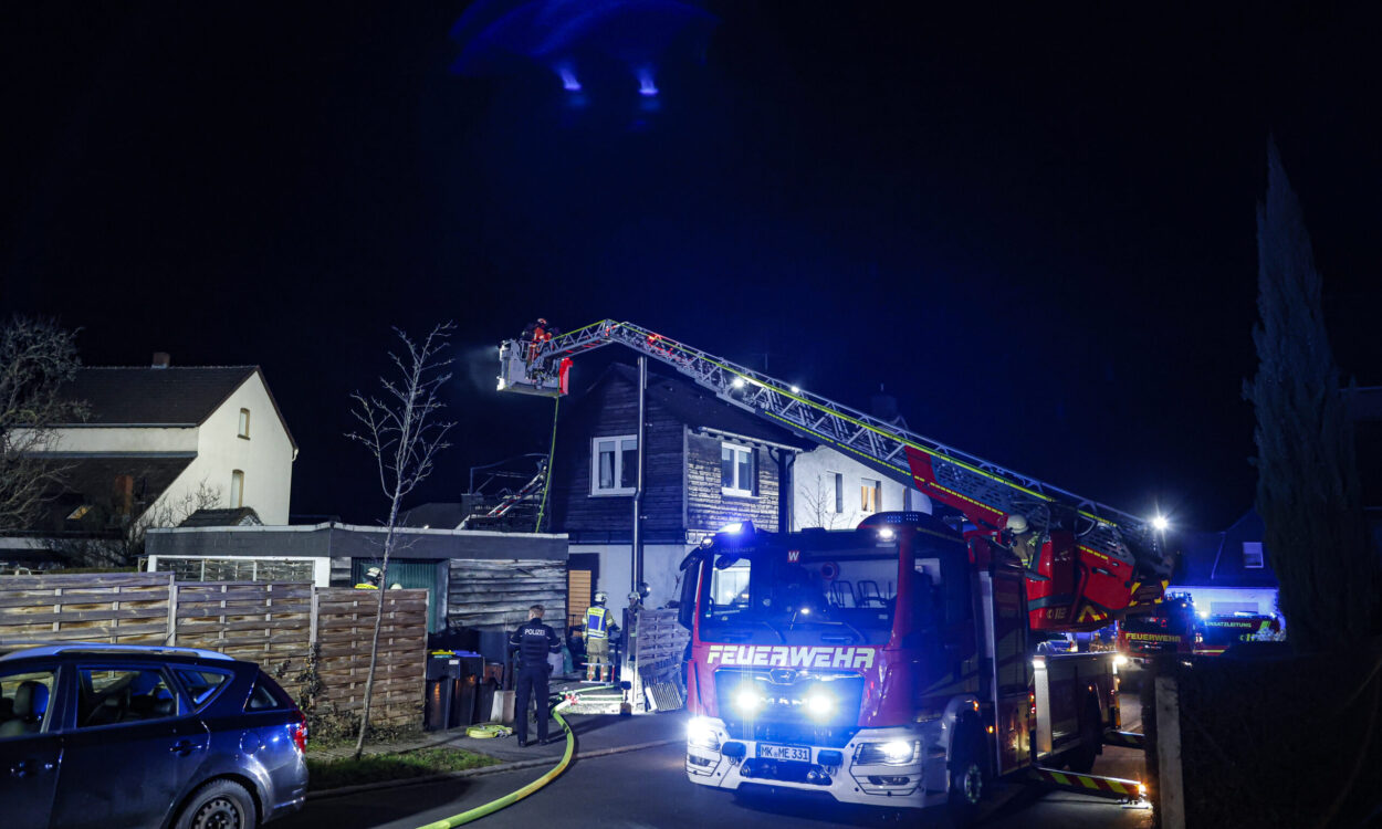 Ein Feuerwehreinsatz im Dunkeln in einem Wohngebiet: Ein Feuerwehrfahrzeug mit ausgefahrener Drehleiter steht vor einem Haus, mehrere Einsatzkräfte arbeiten unter blauem Licht. Schläuche liegen auf der Straße, umliegende Gebäude und parkende Autos sind zu sehen.