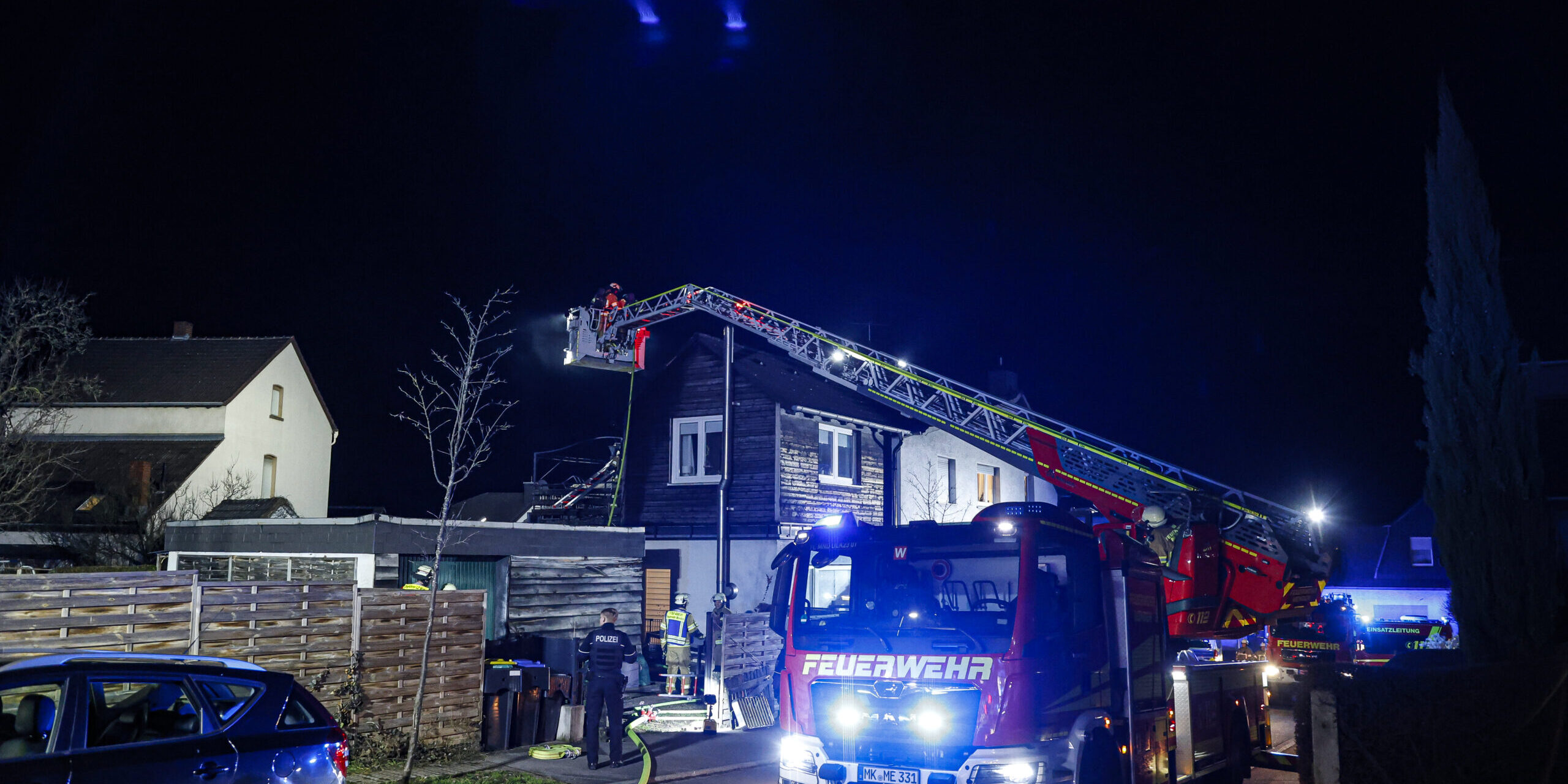 Ein Feuerwehreinsatz im Dunkeln in einem Wohngebiet: Ein Feuerwehrfahrzeug mit ausgefahrener Drehleiter steht vor einem Haus, mehrere Einsatzkräfte arbeiten unter blauem Licht. Schläuche liegen auf der Straße, umliegende Gebäude und parkende Autos sind zu sehen.