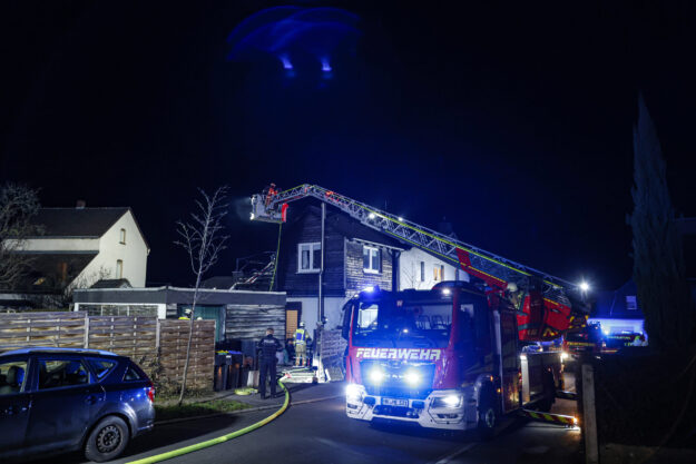 Ein Feuerwehreinsatz im Dunkeln in einem Wohngebiet: Ein Feuerwehrfahrzeug mit ausgefahrener Drehleiter steht vor einem Haus, mehrere Einsatzkräfte arbeiten unter blauem Licht. Schläuche liegen auf der Straße, umliegende Gebäude und parkende Autos sind zu sehen.