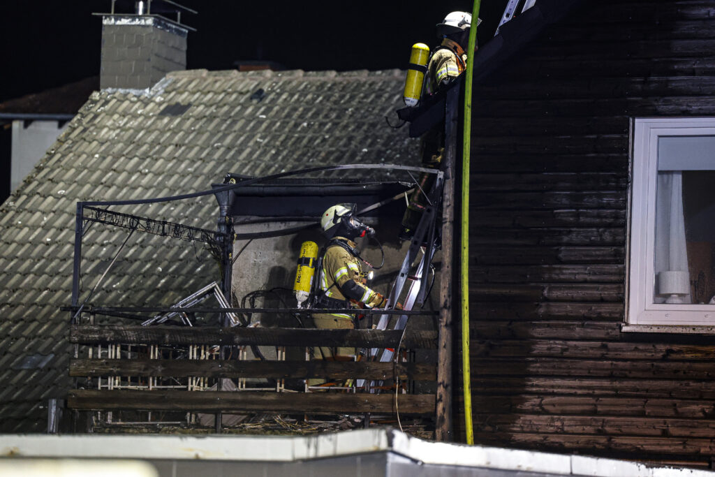Zwei Feuerwehrleute in Schutzkleidung arbeiten nachts an einem beschädigten Balkon eines Wohnhauses. Einer steht auf einer Leiter, der andere auf dem Balkon, beide tragen Atemschutzgeräte; die Fassade ist verrußt und beschädigt.
