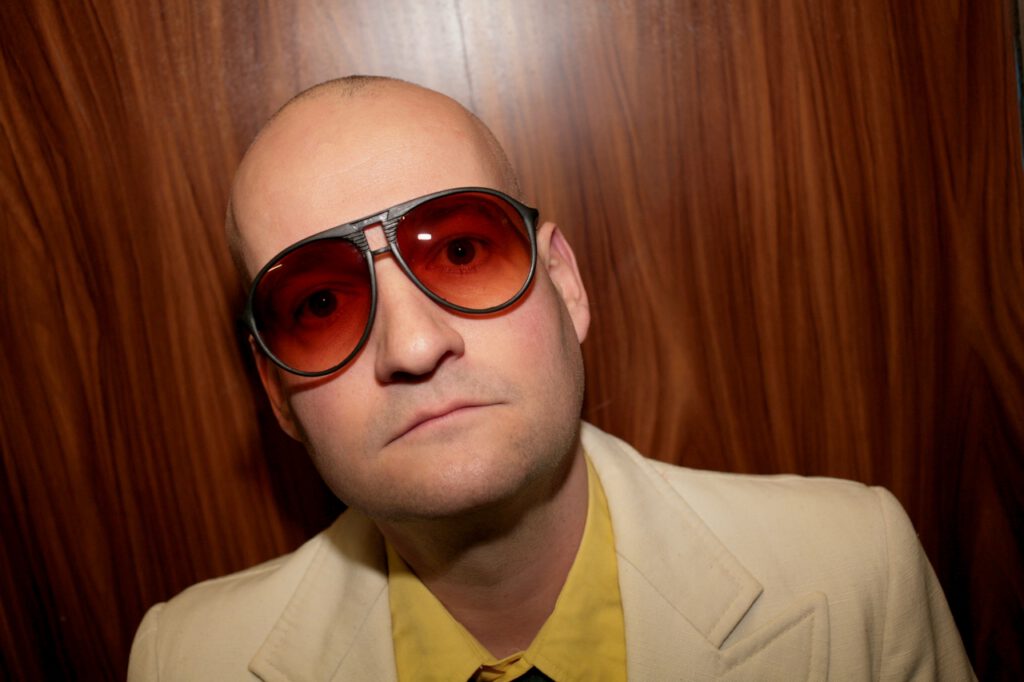 Ein Mann mit rasiertem Kopf und rot getönten Sonnenbrillen blickt direkt in die Kamera. Er trägt ein helles Jackett über einem gelben Hemd, der Hintergrund besteht aus einer holzverkleideten Wand.