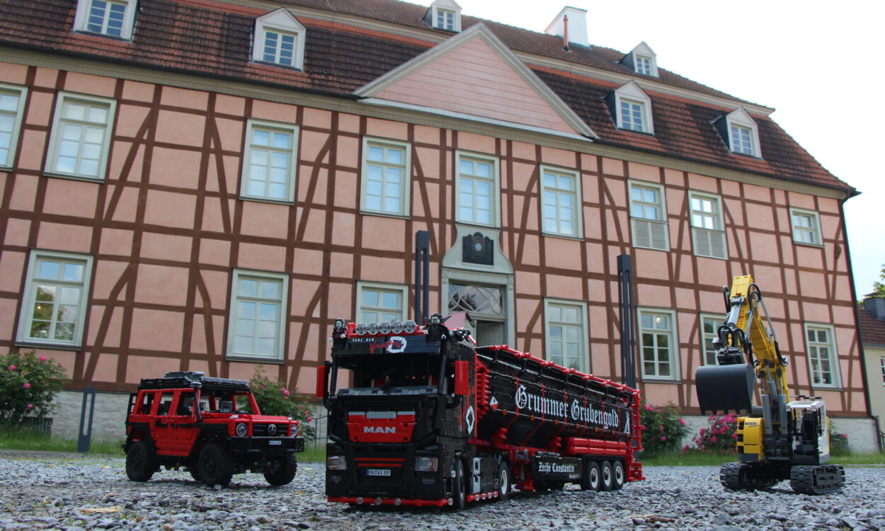 Detailreiche Modellfahrzeuge aus Klemmbausteinen – ein Lkw, ein Geländewagen und ein Bagger – stehen auf Kies vor einem historischen Fachwerkgebäude.