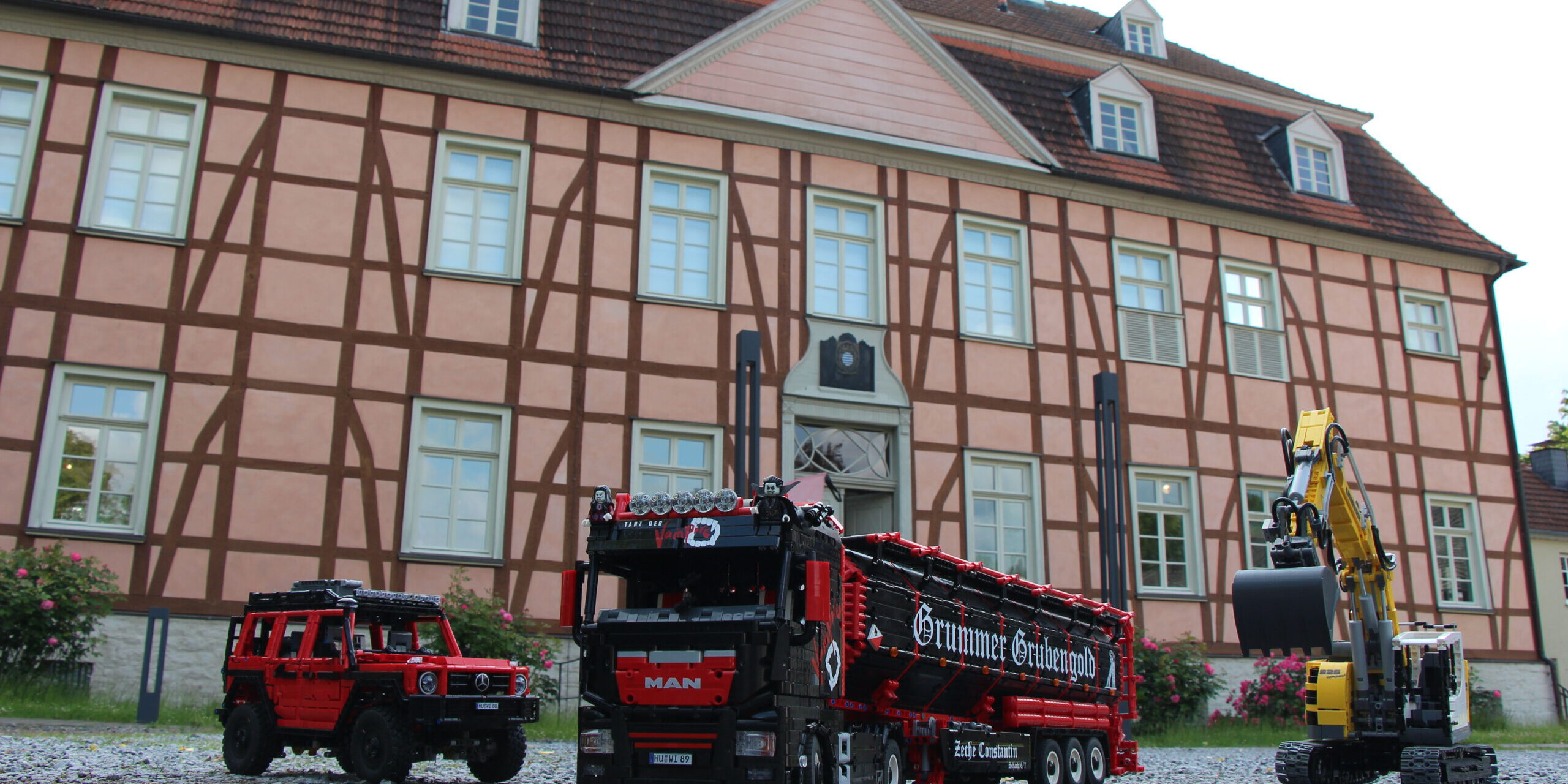Detailreiche Modellfahrzeuge aus Klemmbausteinen – ein Lkw, ein Geländewagen und ein Bagger – stehen auf Kies vor einem historischen Fachwerkgebäude.