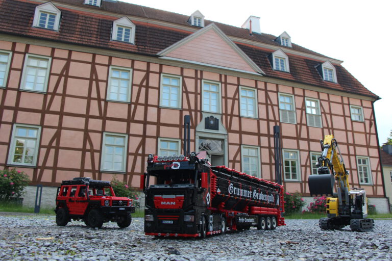 Detailreiche Modellfahrzeuge aus Klemmbausteinen – ein Lkw, ein Geländewagen und ein Bagger – stehen auf Kies vor einem historischen Fachwerkgebäude.